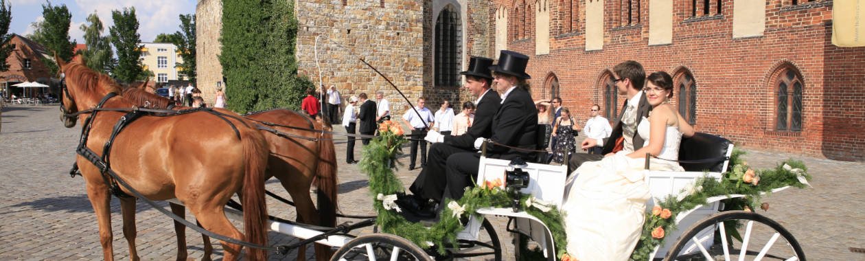 Brautpaar mit Kutscher im Zylinder in offener Hochzeitskutsche mit braunen Pferden vor dem Backsteindom in der Altstadt von Havelberg