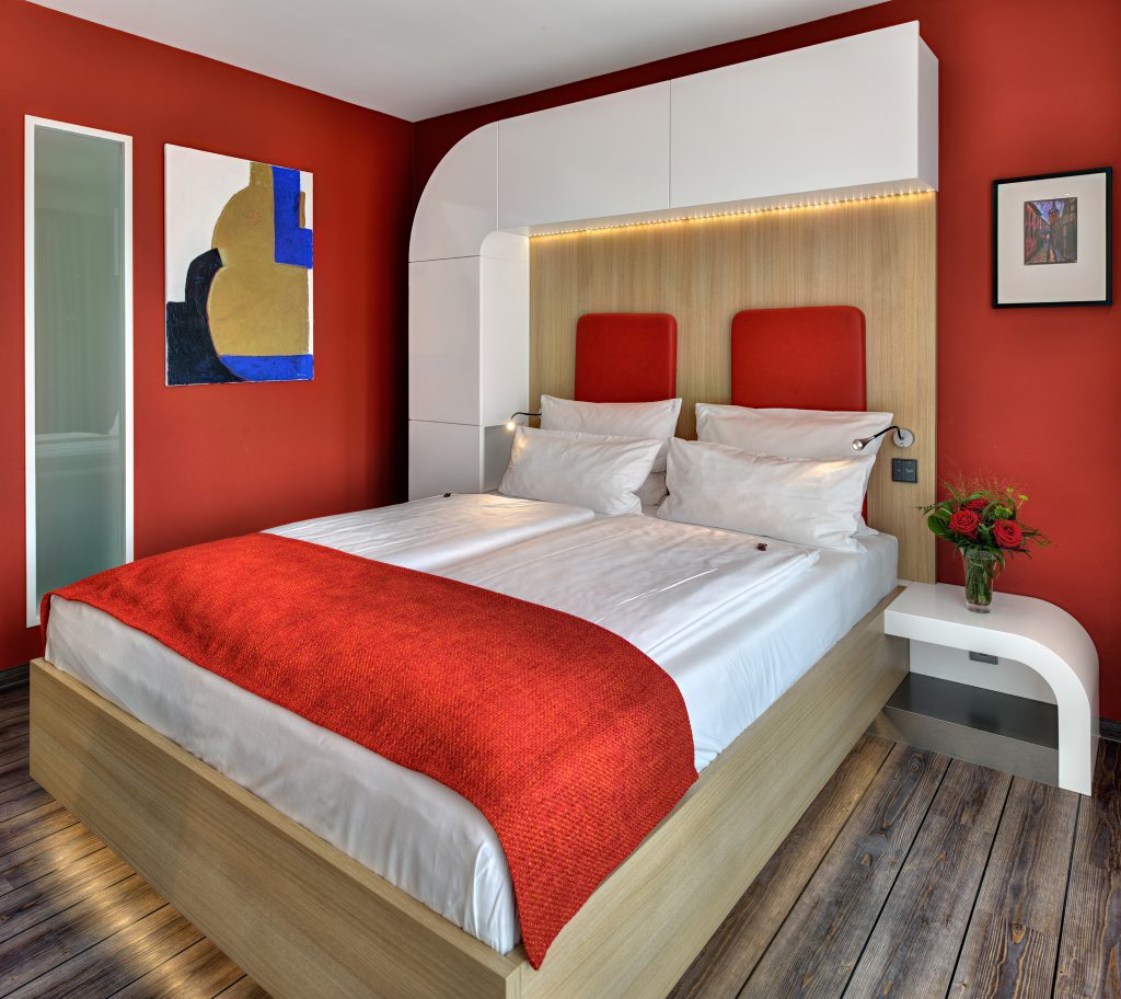 Modernes Doppelzimmer im ArtHotel Kiebitzberg mit roter Wand, Doppelbett, rotem Bettläufer und abstrakter Kunst