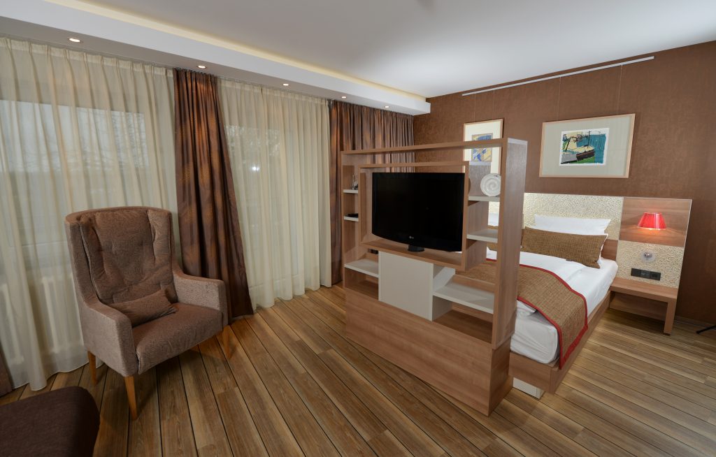 Schlafbereich einer Suite im ArtHotel Kiebitzberg mit Doppelbett, Holz-Raumteiler mit TV und Ohrensessel am Fenster