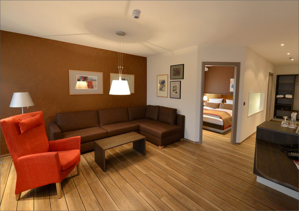 Wohnzimmer einer Suite im ArtHotel Kiebitzberg mit brauner Eckcouch, orangem Sessel und Blick ins Schlafzimmer