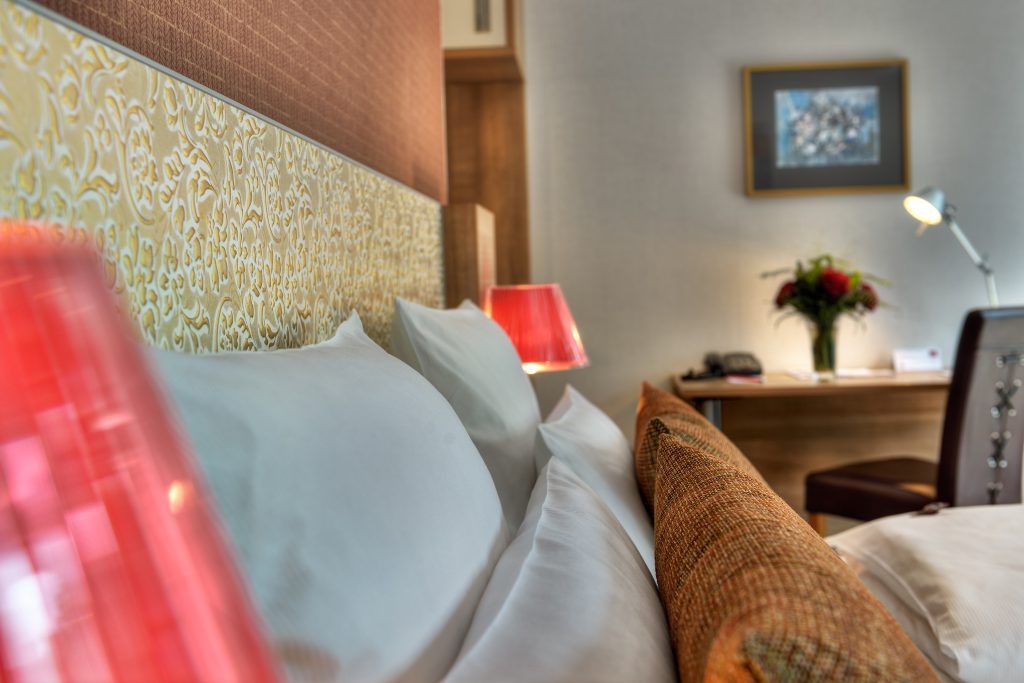 Detail des Comfort-Zimmers im ArtHotel Kiebitzberg: weiße Kissen, ornamentale Goldtapete und rote Lampe am Bett