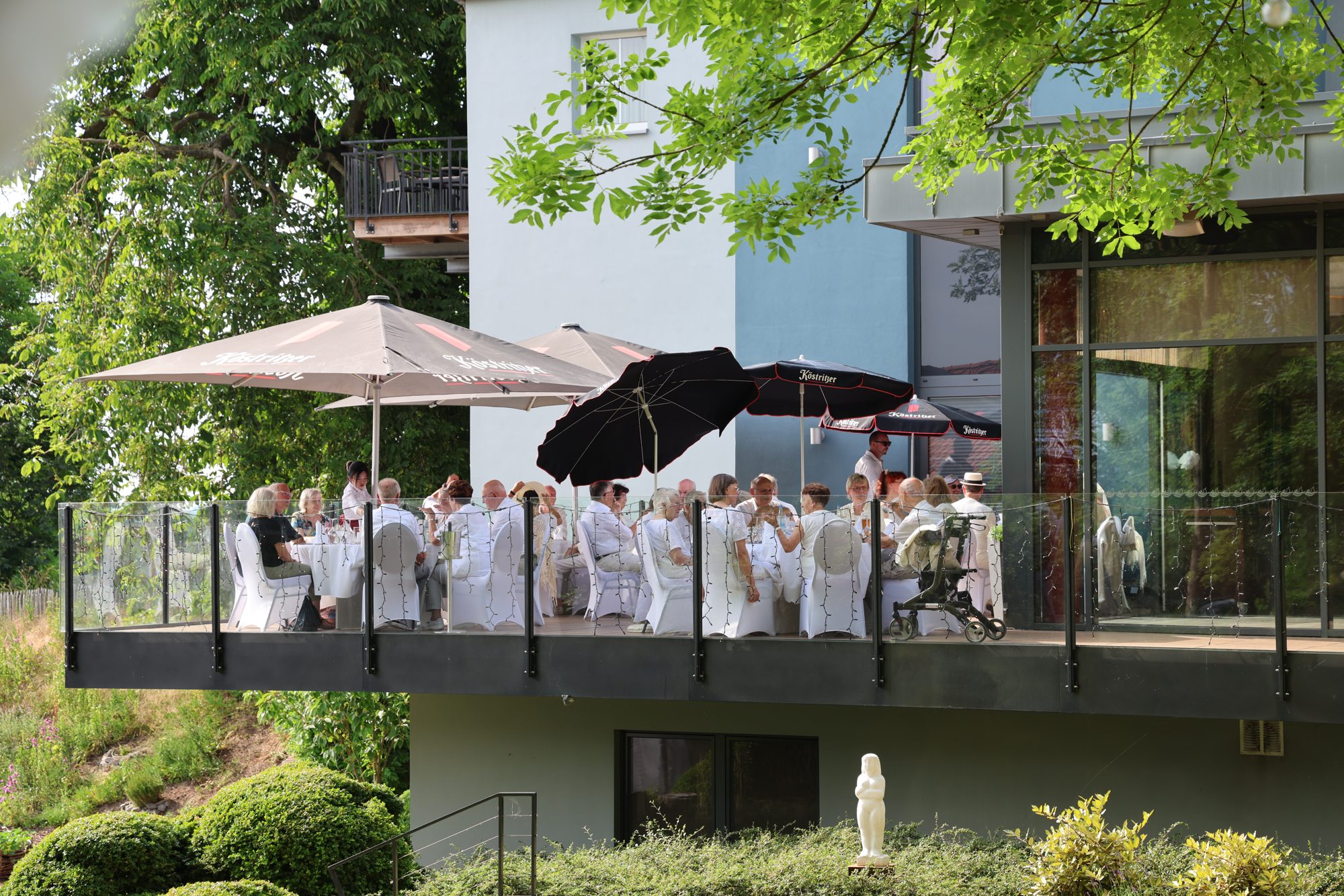 Festgesellschaft in weißer Kleidung feiert auf der Außenterrasse des ArtHotel Kiebitzberg unter Sonnenschirmen
