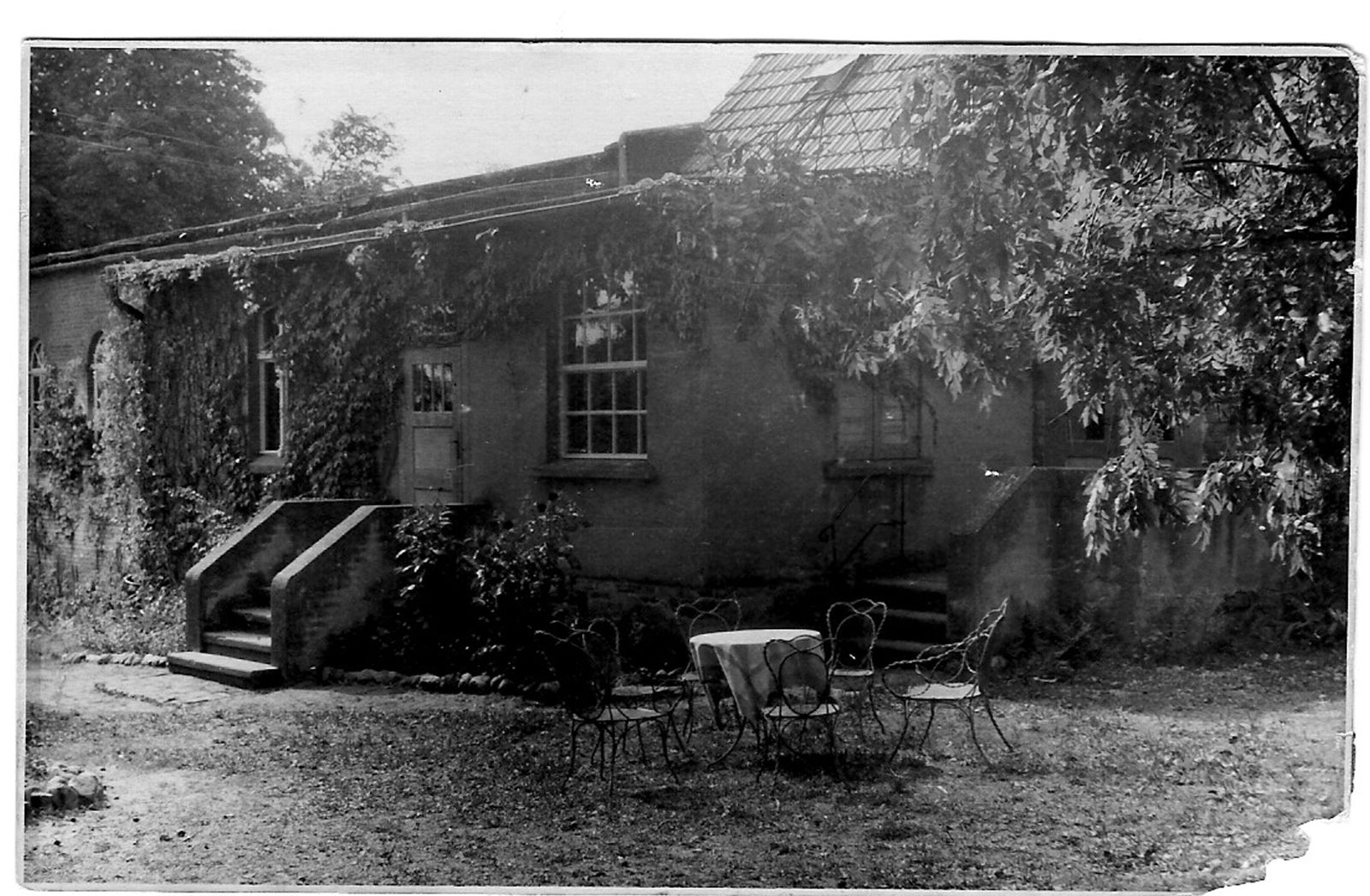 Historisches Schwarzweiß-Foto eines efeubewachsenen Gartenhauses mit Glasdach, Freitreppe und Bistrotisch im Garten