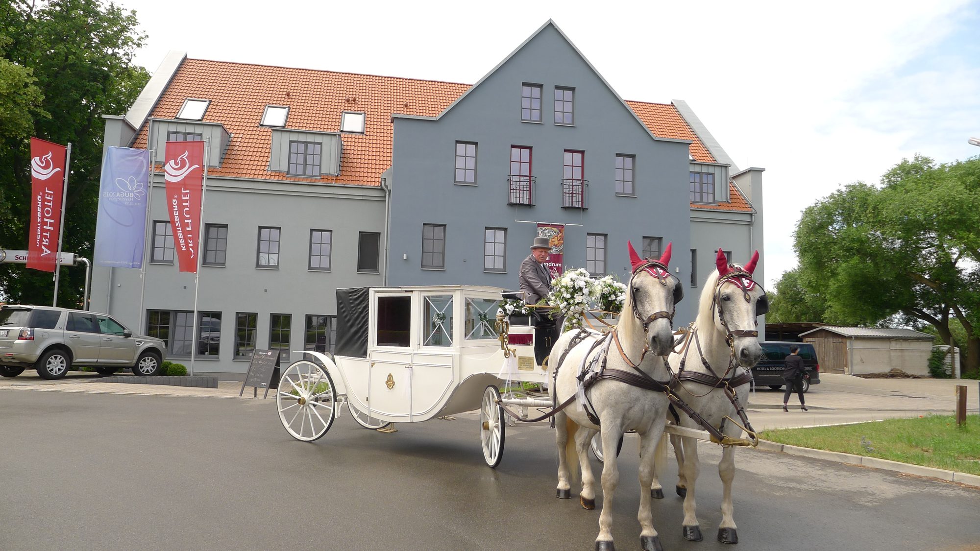 Weiße Hochzeitskutsche mit zwei Schimmeln und Kutscher vor dem ArtHotel Kiebitzberg mit ArtHotel-Fahnen