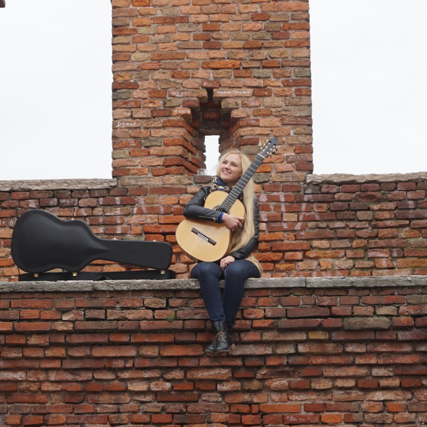 Gitarristin Julia Trintschuk sitzt mit klassischer Konzertgitarre auf einer historischen Backsteinmauer, daneben ihr Gitarrenkoffer