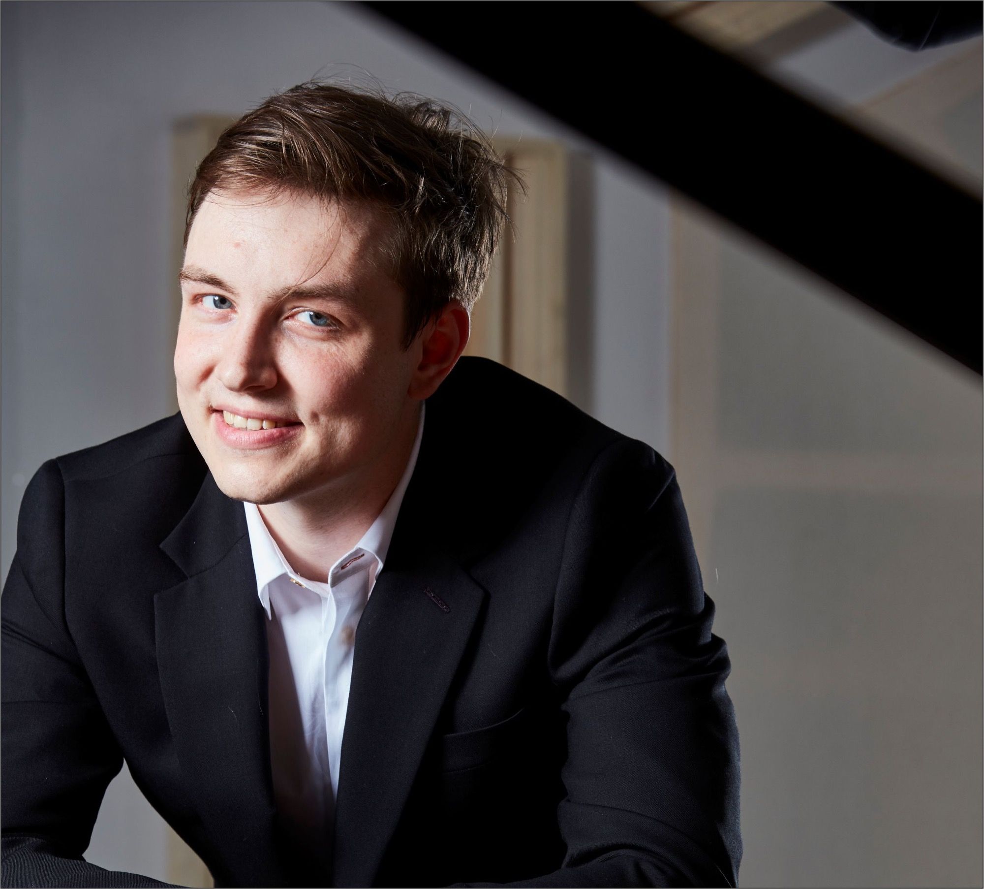 Pianist Florian Heinisch im dunklen Anzug lächelt an einen schwarzen Flügel gelehnt – Künstlerporträt zur Konzertreihe im ArtHotel Kiebitzberg