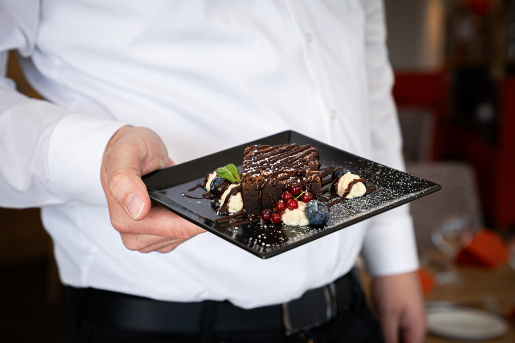 Servicekraft im weißen Hemd serviert Schokoladen-Dessert mit Beeren und Sahne auf schwarzer Platte im Restaurant Schmokenberg