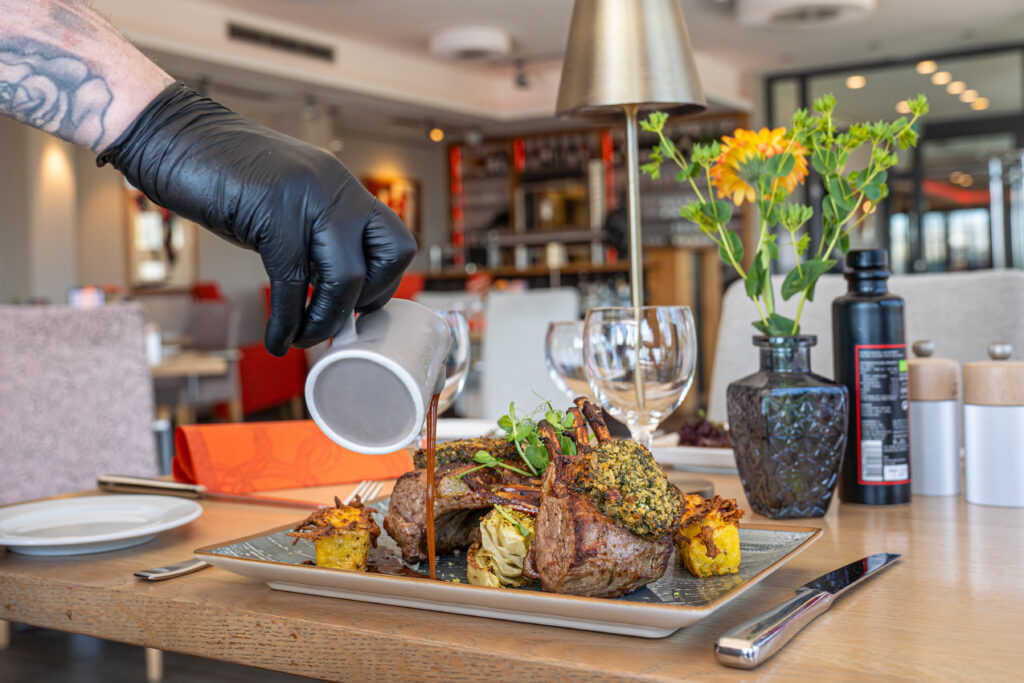 Lammkarree wird am Gasttisch im Restaurant Schmokenberg mit Sauce angerichtet, Weingläser und Blumen im Hintergrund