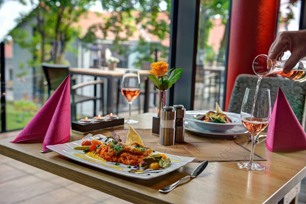 Gedeckter Tisch am Panoramafenster des Restaurants Schmokenberg mit Hauptgericht und eingeschenktem Roséwein, Blick ins Grüne