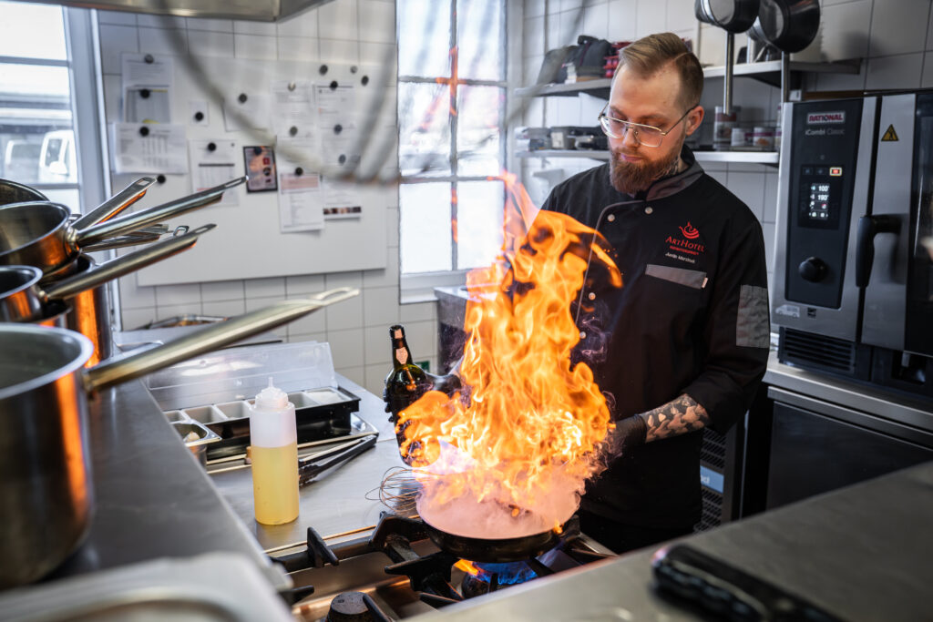 Koch in schwarzer ArtHotel-Kochjacke flambiert in der Küche eine Pfanne mit hoch aufschlagender Flamme